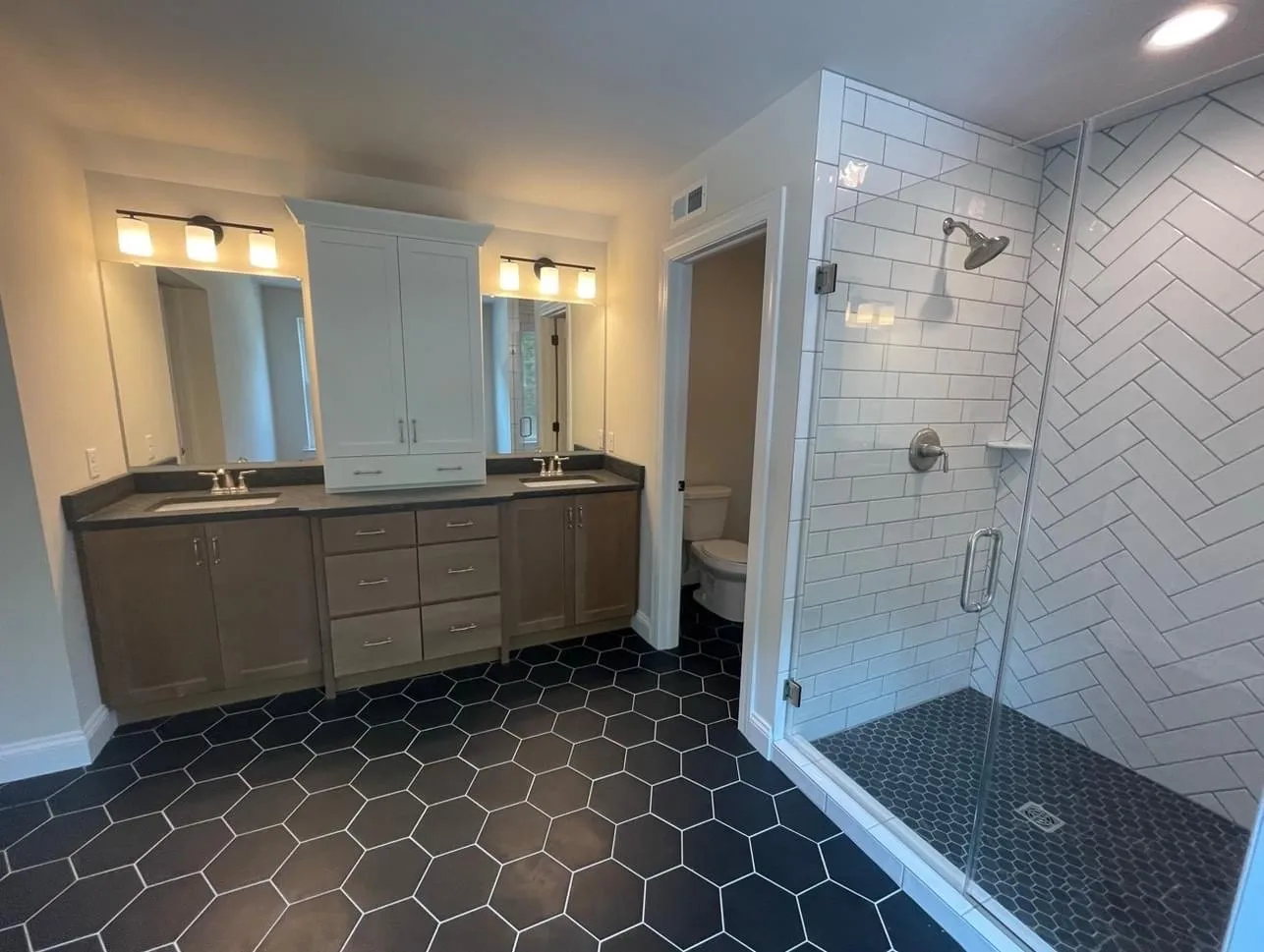 A bathroom vanity featuring a central white cabinet tower between two sinks with dark countertops.