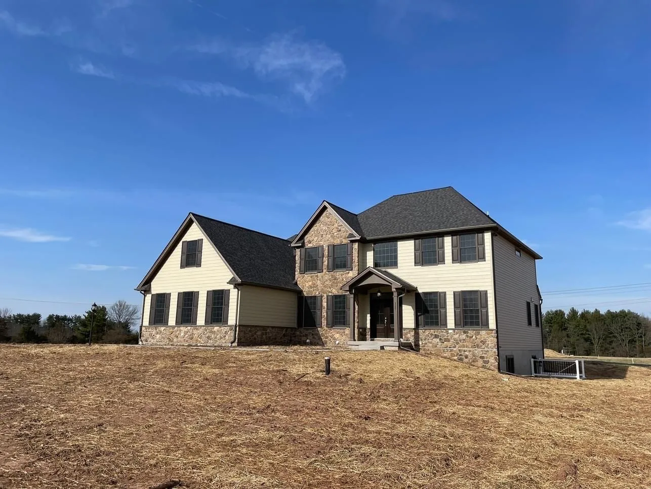A two-story residential home exterior featuring tan siding and stone veneer accents under a blue sky.