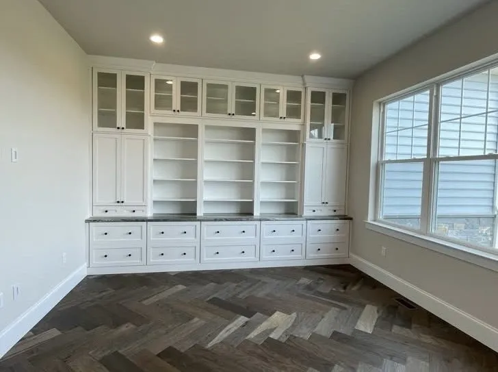 A large white built-in bookcase with glass doors and drawers stands against a wall with herringbone flooring.