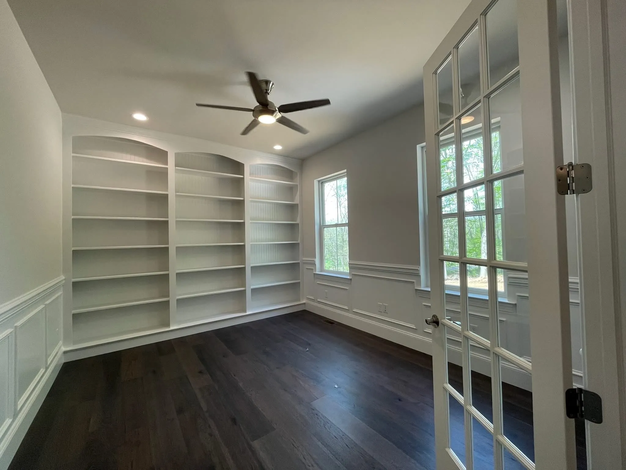White built-in bookshelves with arched tops installed in a home office with wainscoting and hardwood floors.
