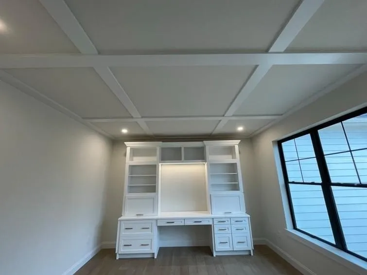 A white built-in desk and shelving unit sits beneath a white coffered ceiling in an office.