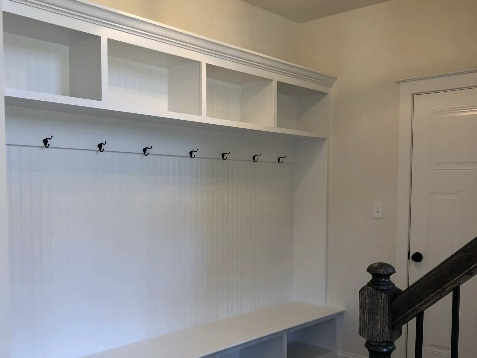 White mudroom lockers with beadboard backing and black hooks installed next to a wooden staircase.